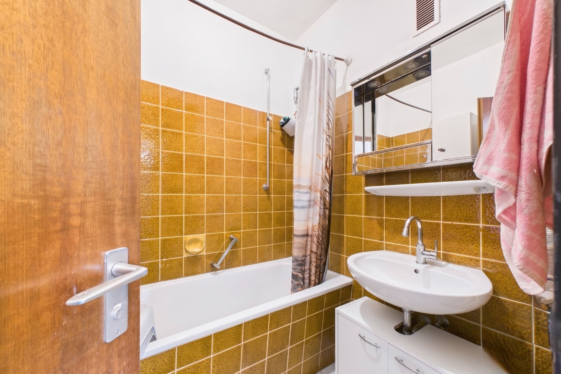 Kleines Bad mit braunen Fliesen, Badewanne, Waschbecken und Spiegelschrank.