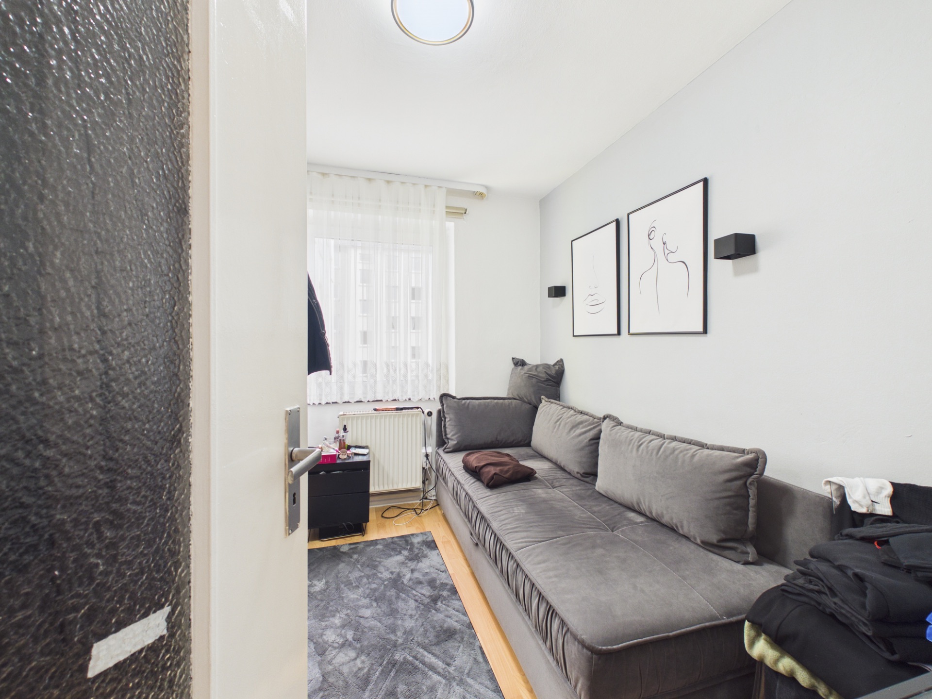 Kleines Wohnzimmer mit grauem Sofa, Wandbildern und Fenster mit Vorhang.
