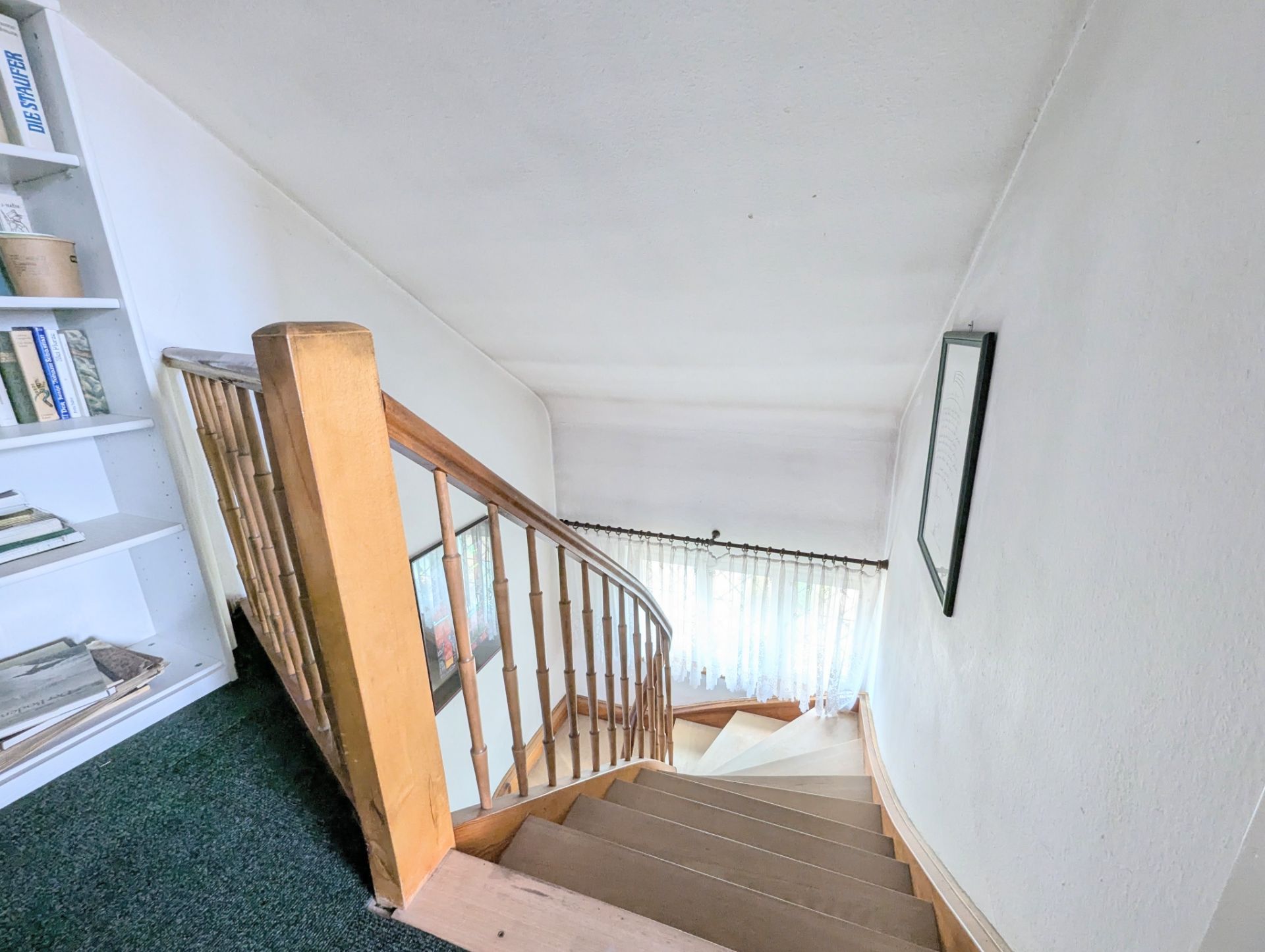 Holztreppe mit Geländer, Fenster mit Vorhang am unteren Ende, Bücherregal links.