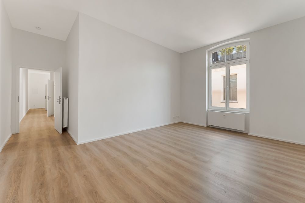 Leerer, heller Raum mit großem Fenster und Holzboden, Tür führt in einen Flur.