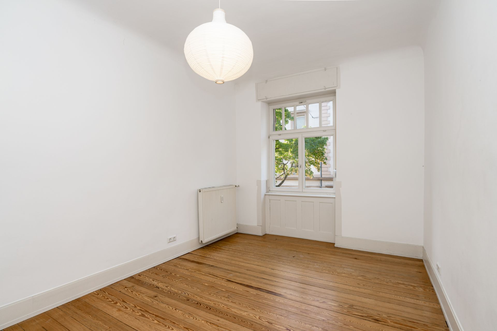 Leerer Raum mit Holzboden, weißer Wand, Fenster, Heizung und Deckenlampe.