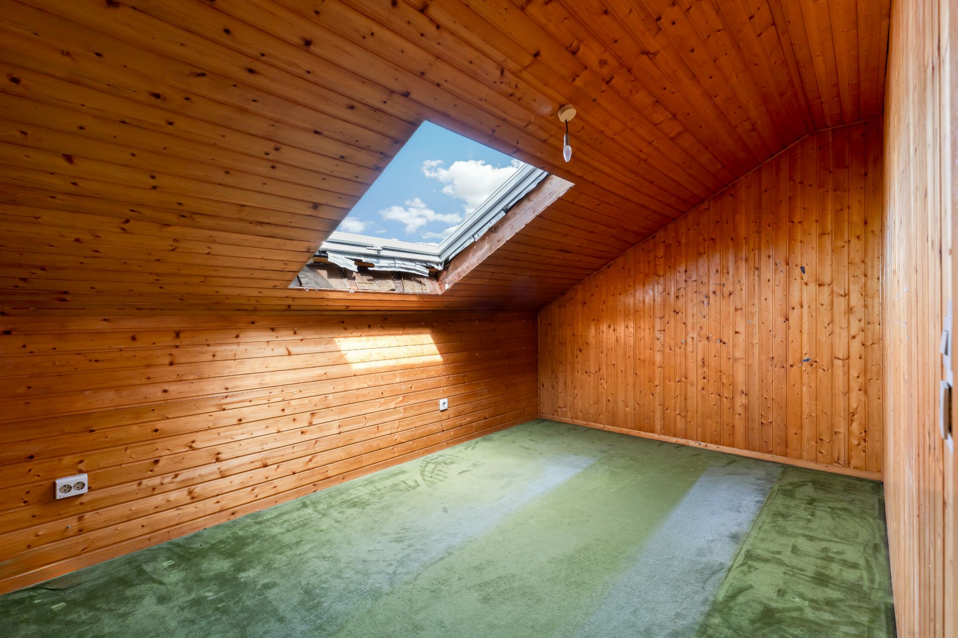 Dachzimmer mit Holzverkleidung, grünem Teppich und großem Dachfenster.