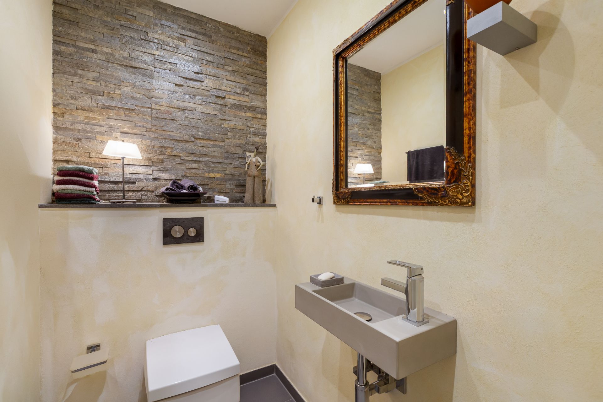 Modernes Gästebad mit Steinwand, Spiegel, Toilette und kleinem Waschbecken.