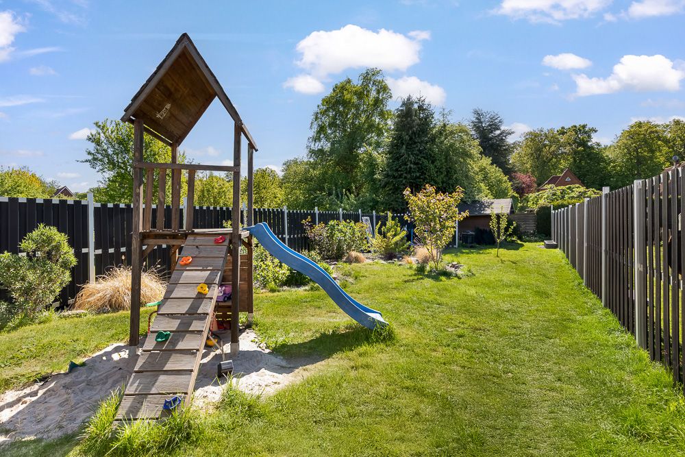 Holzspielturm mit Rutsche und Sandkasten im Garten mit grünem Rasen und Zaun.