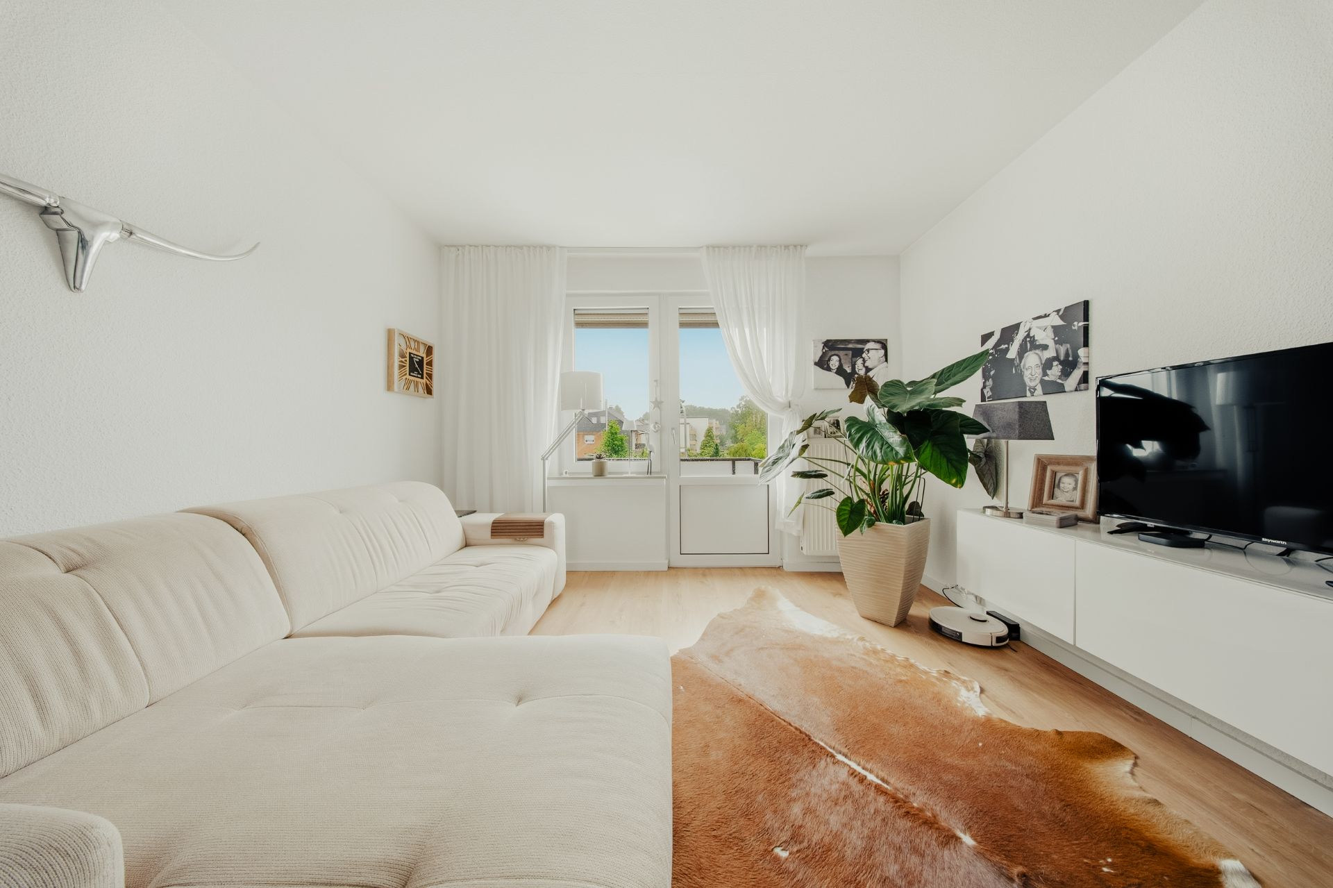 Helles Wohnzimmer mit beigem Sofa, Teppich, TV, großer Pflanze und Fenster mit Blick nach draußen.