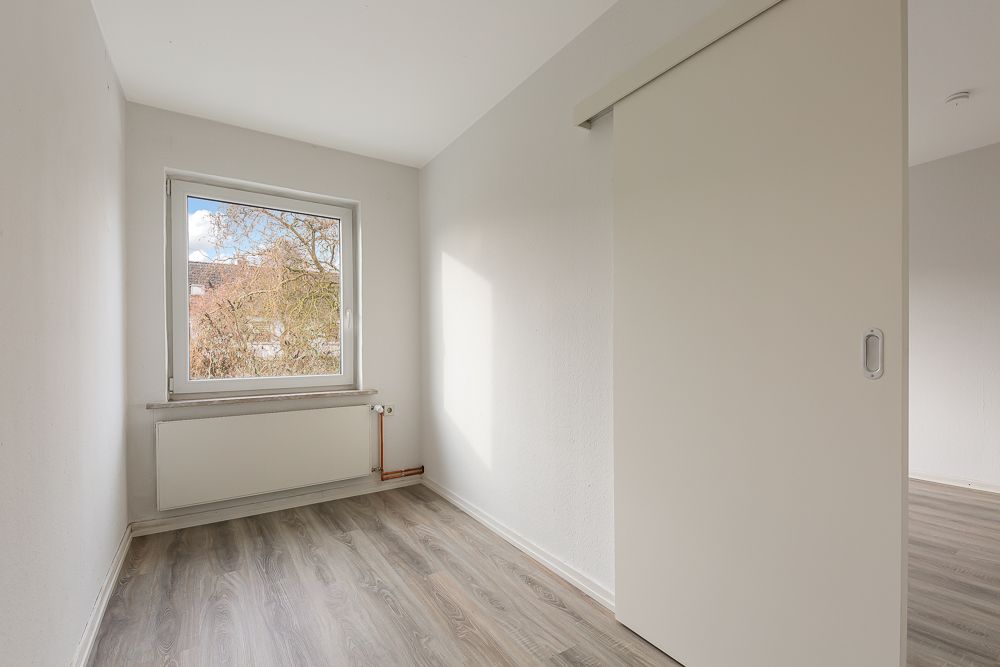 Kleiner, leerer Raum mit Fenster, Heizkörper und Schiebetür, heller Boden.
