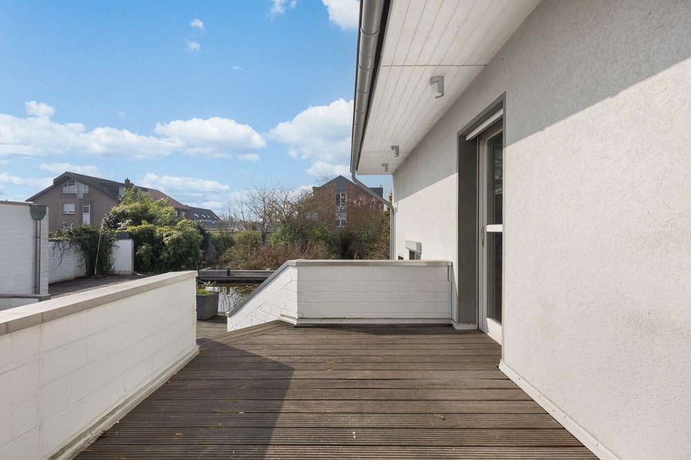 Große Holzterrasse mit weißem Geländer an einem Wohnhaus, Blick auf Nachbarhäuser und Bäume.