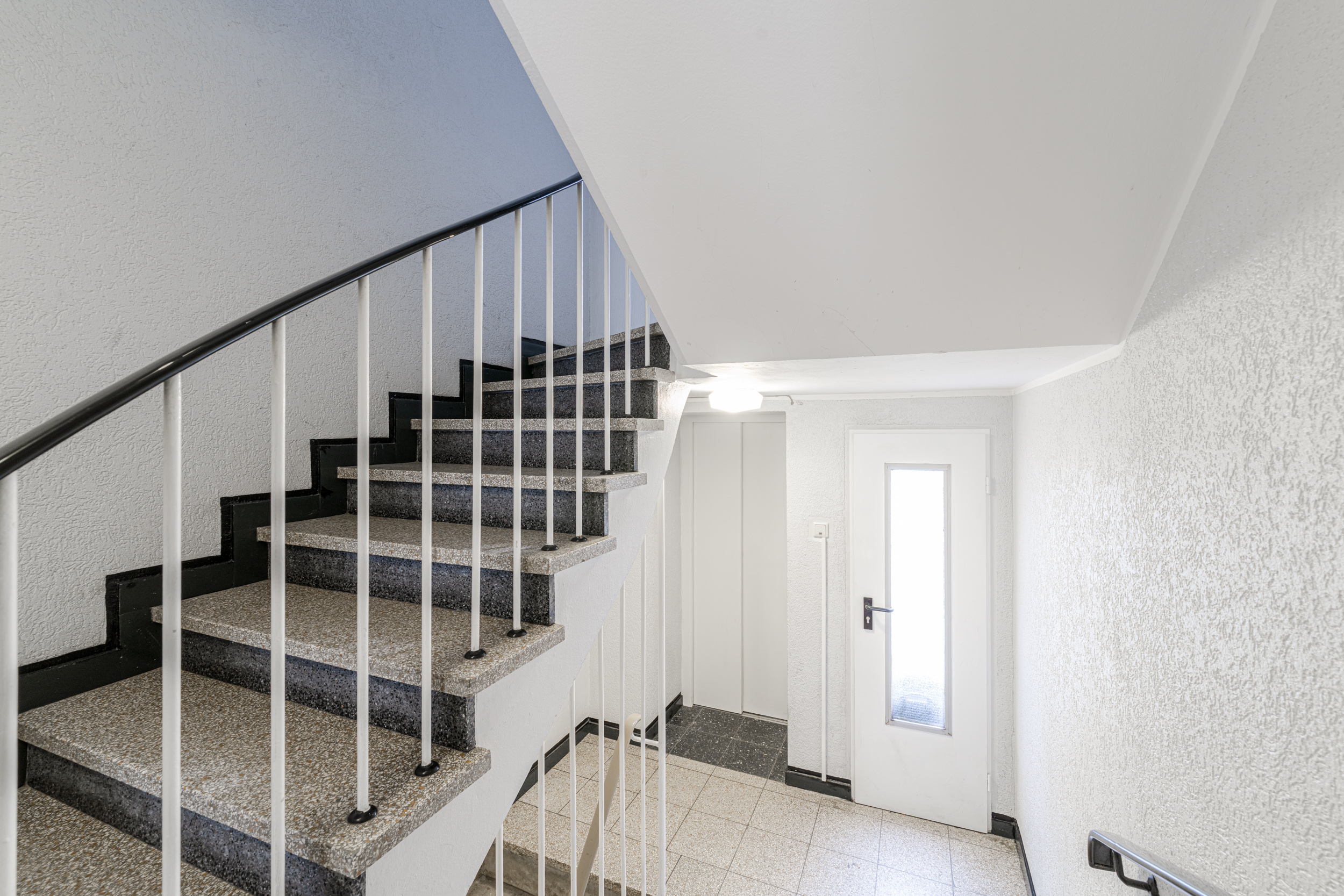 Helles Treppenhaus mit Steintreppe, Handlauf und weißer Tür mit Glaseinsatz.
