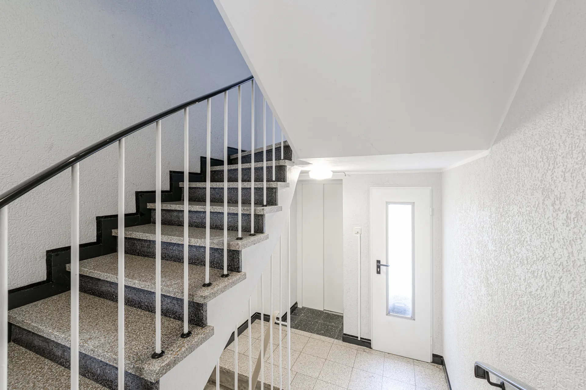 Helles Treppenhaus mit Steintreppe, Handlauf und weißer Tür mit Glaseinsatz.