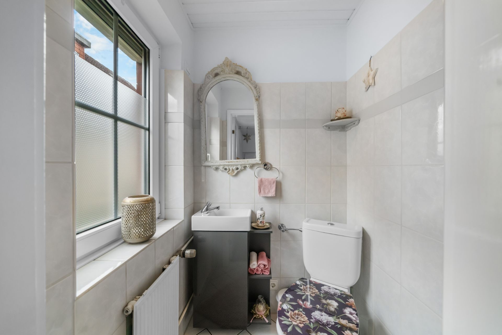 Kleines Badezimmer mit Fenster, Waschbecken, Spiegel und Toilette mit gemustertem Sitz.