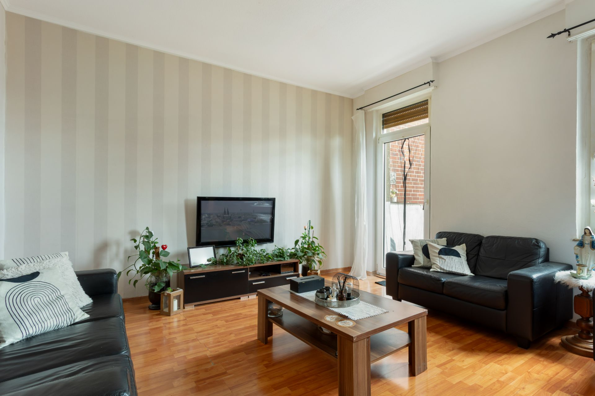 Wohnzimmer mit zwei schwarzen Sofas, Couchtisch, Fernseher und Zimmerpflanzen auf Parkettboden.