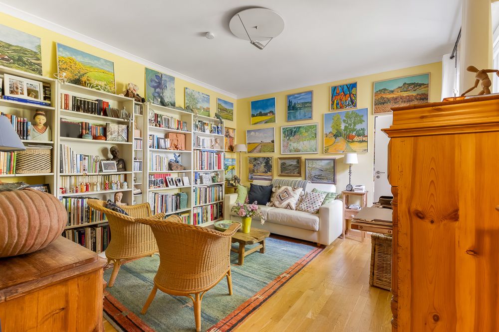 Helles Wohnzimmer mit Sofa, Sesseln, Bücherregalen und vielen Gemälden an gelben Wänden.