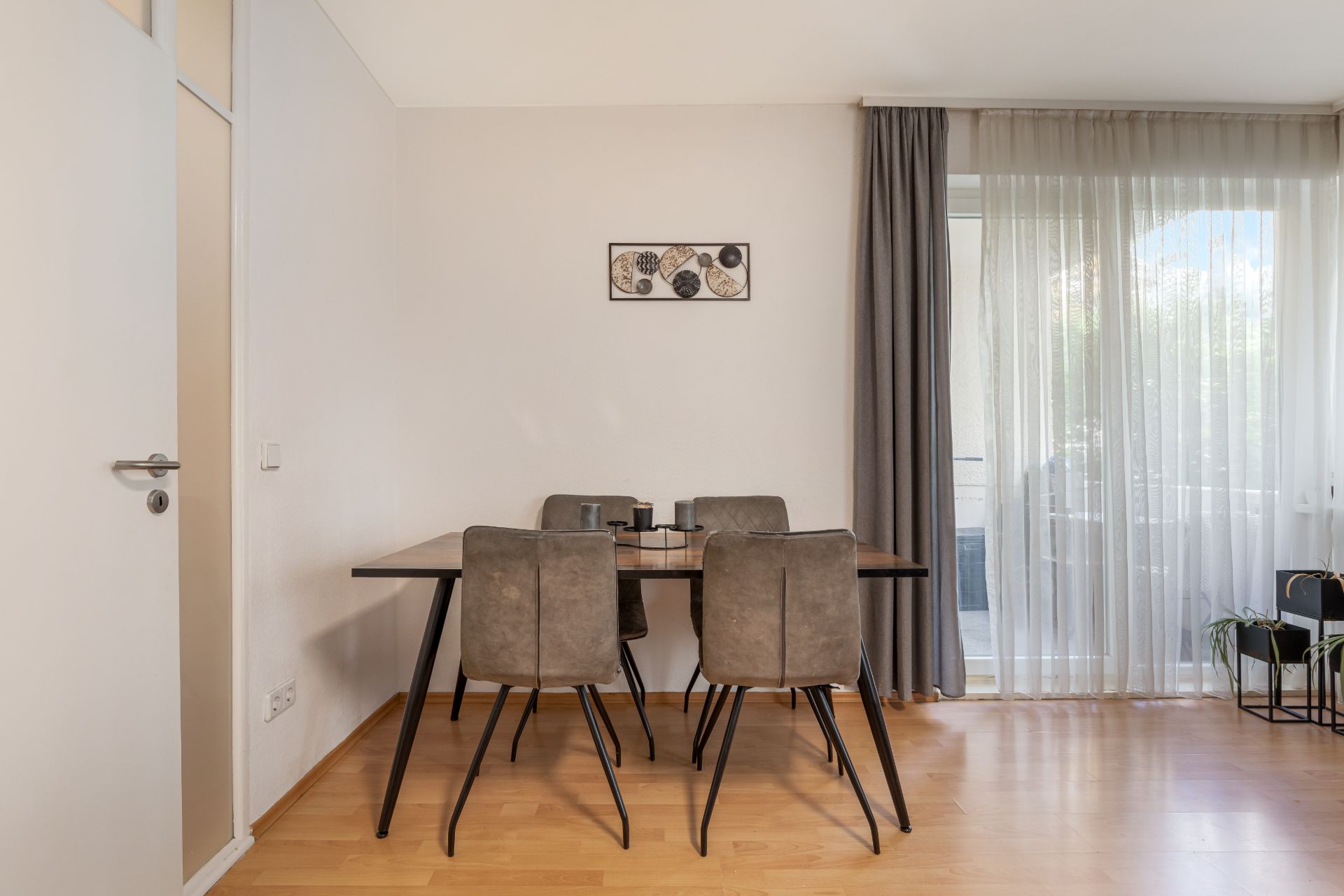 Essbereich mit Holztisch und vier Stühlen vor einer Wand, daneben ein großes Fenster mit Vorhängen.
