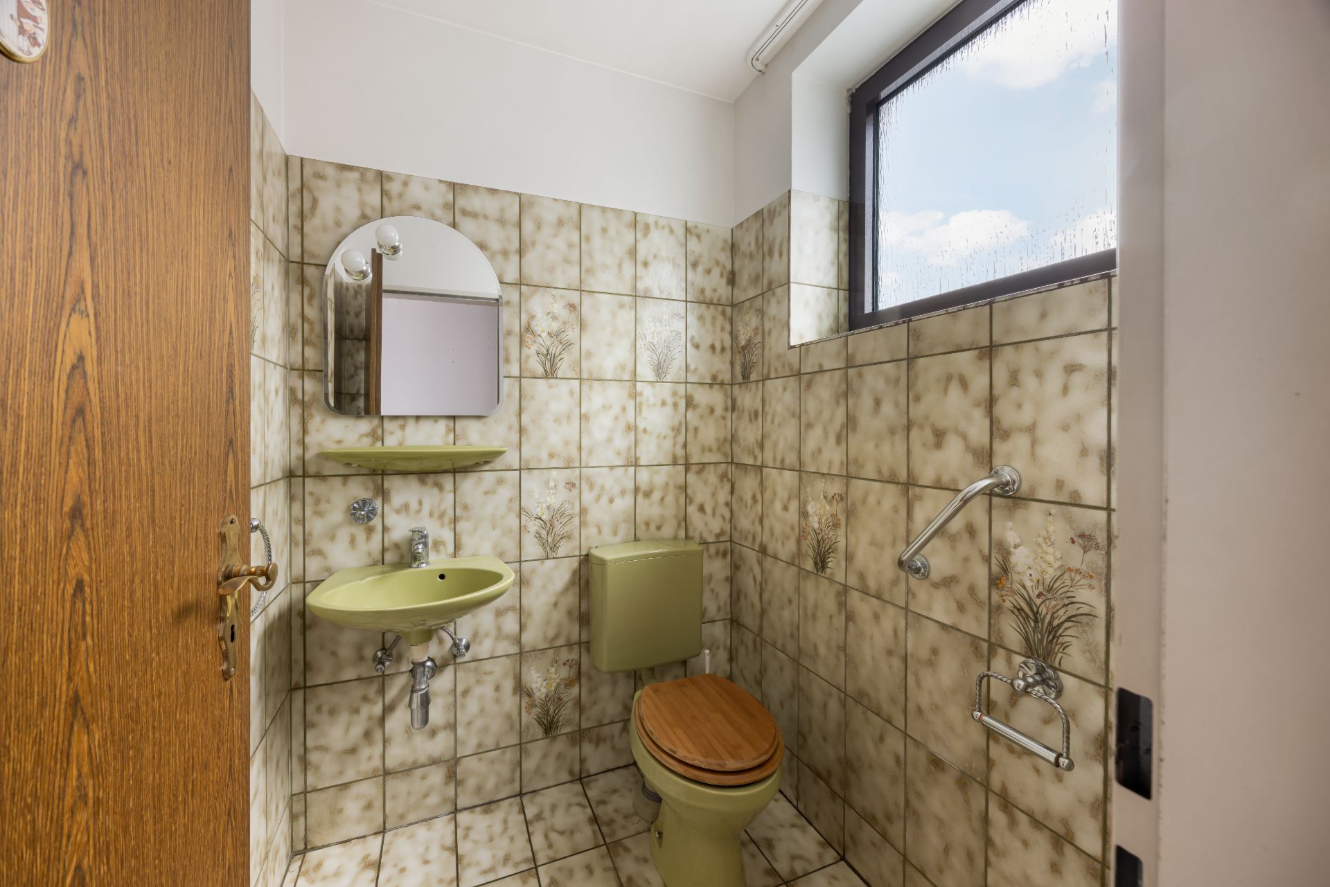 Kleines Badezimmer mit grünem WC, Waschbecken, Spiegel und Fliesen mit Blumenmuster.
