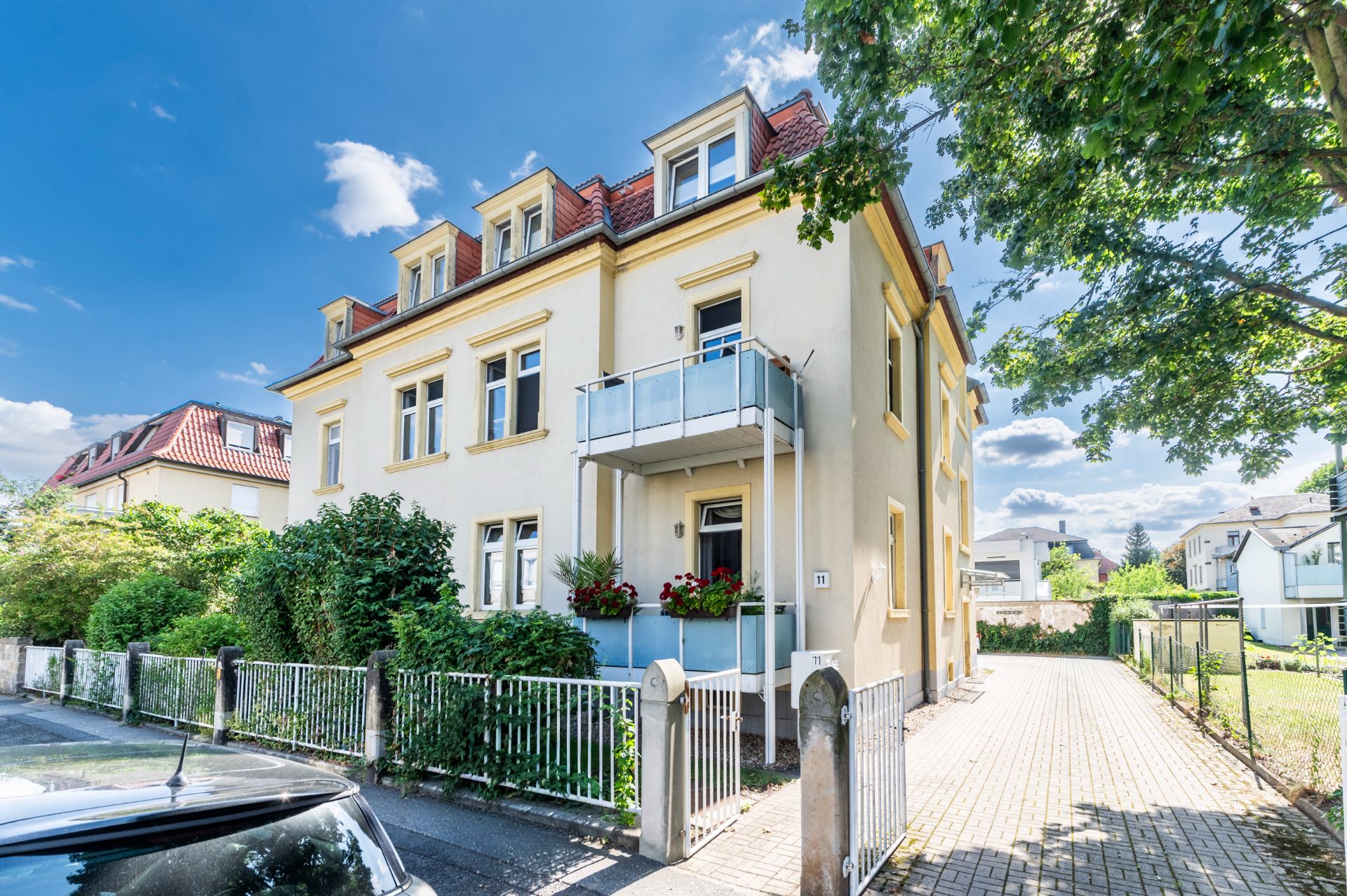 Zweistöckiges Wohnhaus mit Balkon, Vorgarten und Einfahrt, umgeben von Bäumen und Zaun.