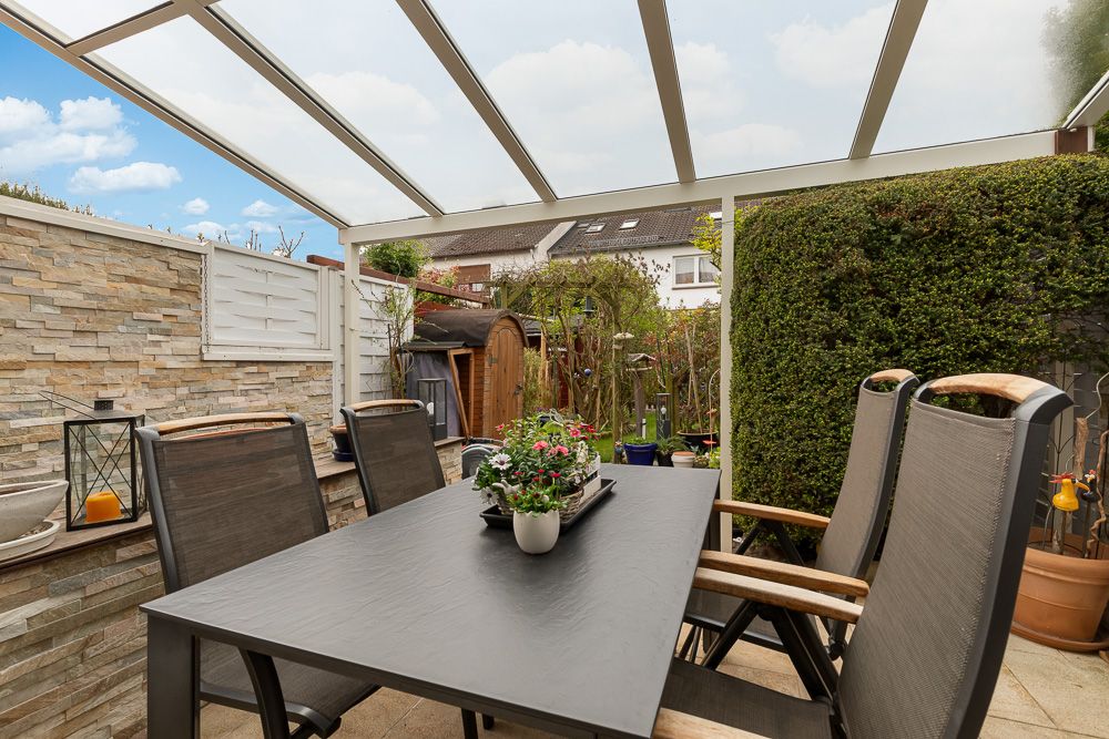 Überdachte Terrasse mit Esstisch, vier Stühlen und Pflanze auf dem Tisch.