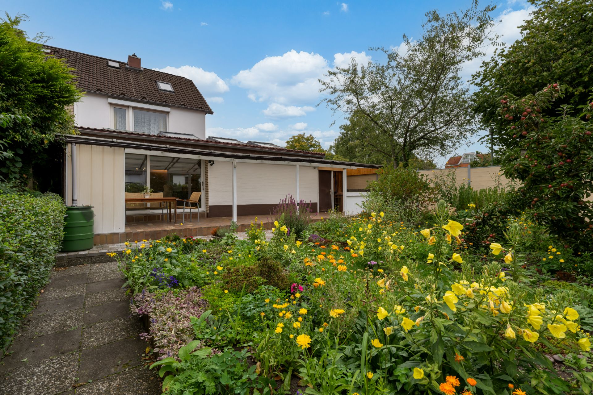 Haus mit großem Garten und vielen bunten Blumen, dahinter Terrasse mit Stühlen.