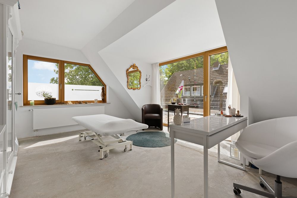 Helles Zimmer mit Behandlungsliege, Schreibtisch, Sesseln und großen Fenstern mit Blick nach draußen.