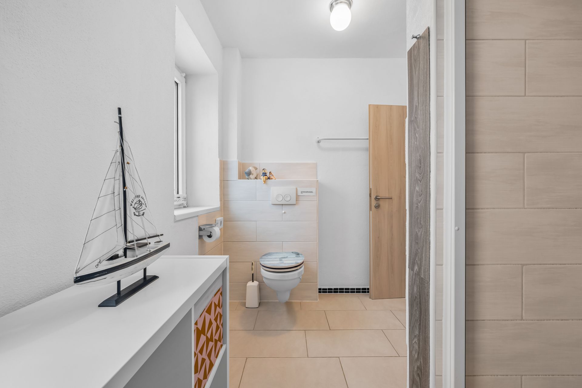 Modernes Badezimmer mit WC, Fenster, kleinem Schrank und Modellsegelboot auf dem Schrank.