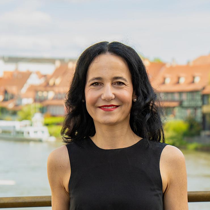 Frau mit dunklen Haaren steht draußen vor einem Fluss und Häusern mit roten Dächern.