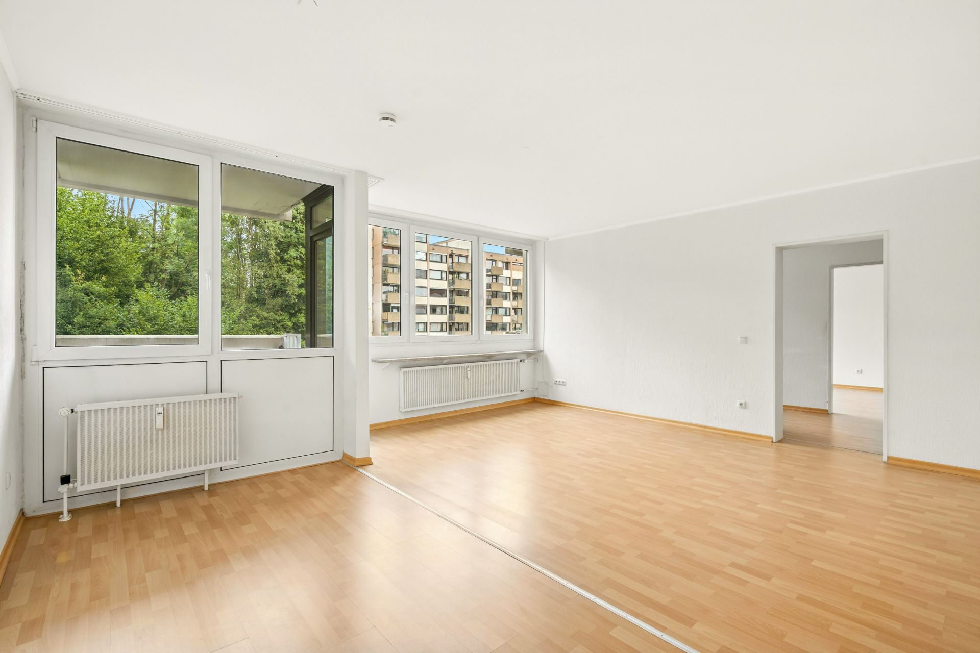Helles Wohnzimmer mit großen Fenstern, Parkettboden und Zugang zu einem weiteren Raum.