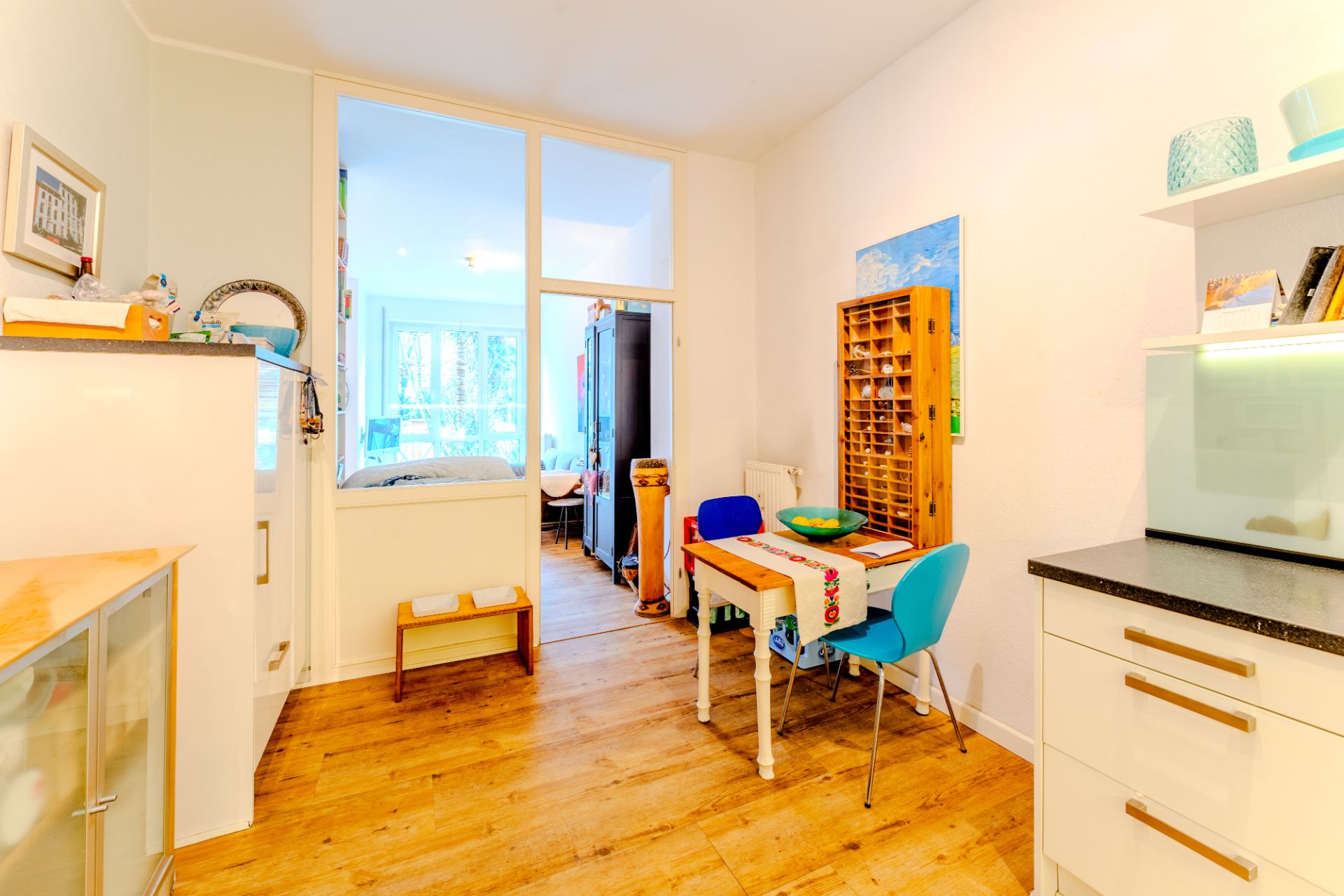 Helle Küche mit Esstisch, zwei Stühlen und Holzboden, Blick ins Wohnzimmer mit großen Fenstern.