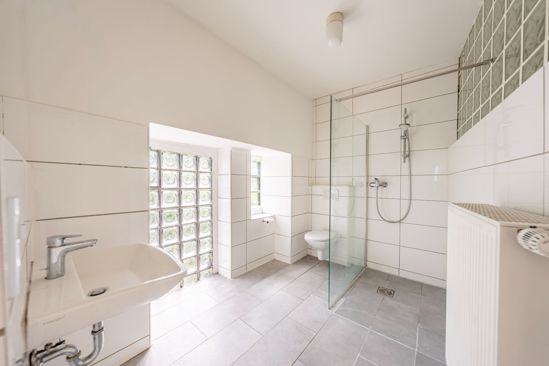 Modernes, weiß gefliestes Badezimmer mit Dusche, Waschbecken, Toilette und Glasbausteinfenster.