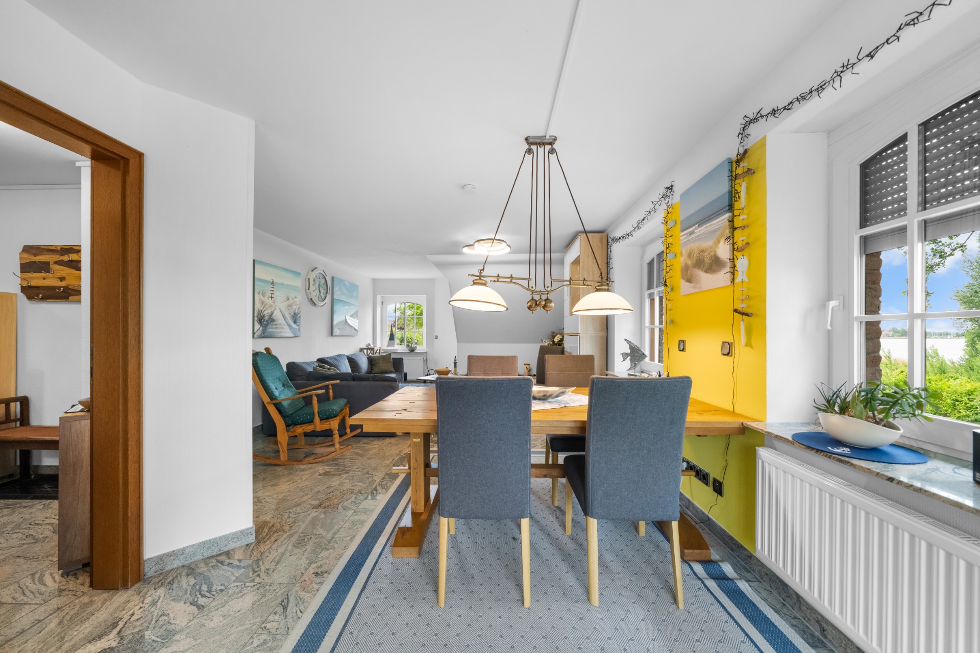 Essbereich mit Holztisch und vier Stühlen, daneben ein Sofa und große Fenster mit Blick ins Grüne.