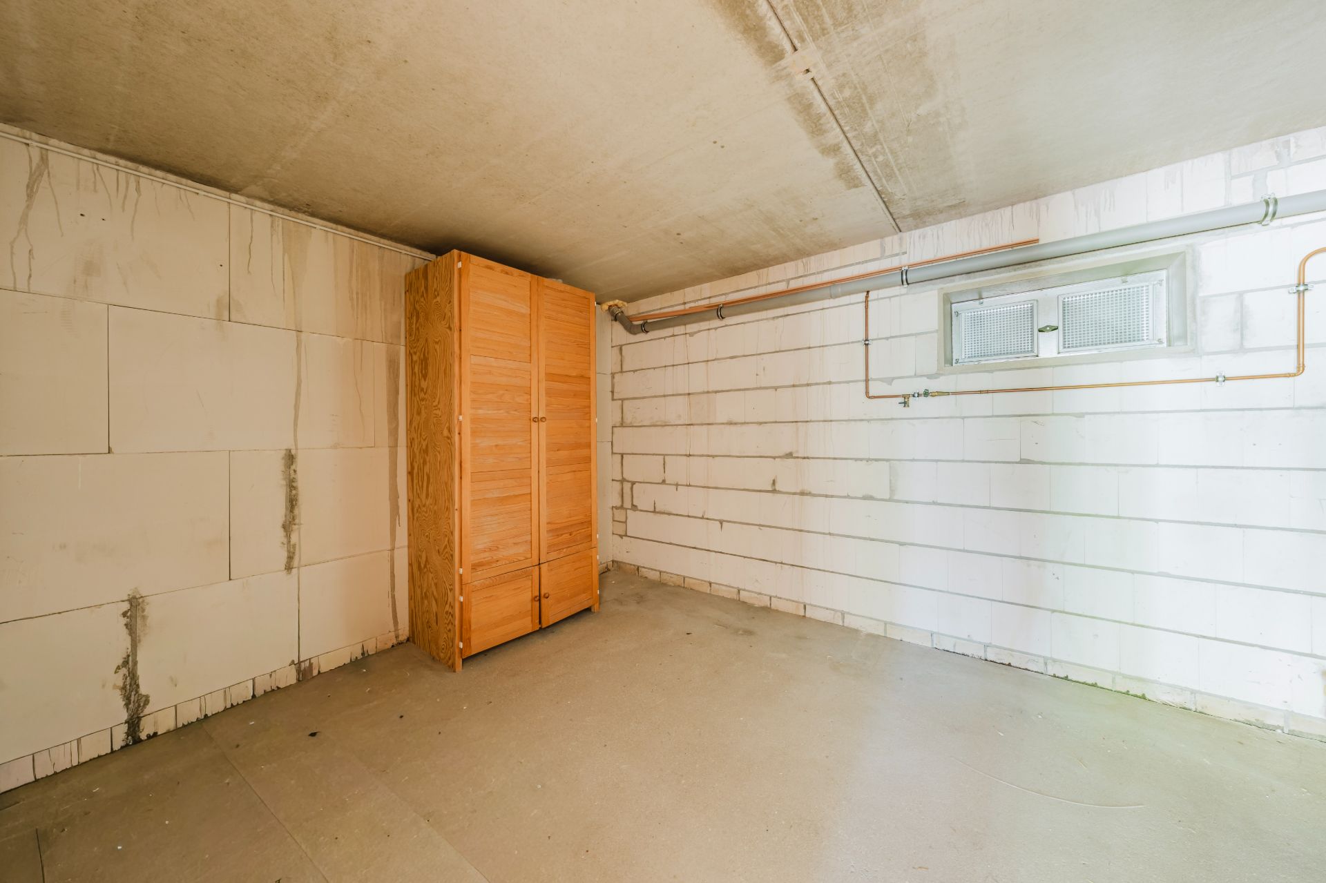 Leerer Kellerraum mit weißem Mauerwerk und Holzschrank an der Wand.