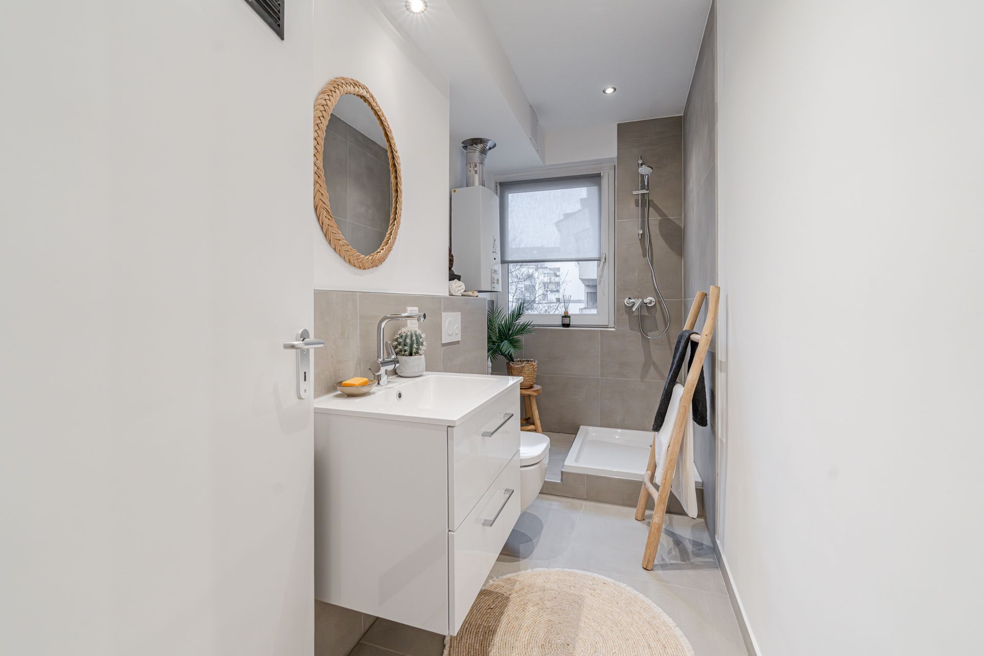 Modernes, helles Badezimmer mit Dusche, Waschbecken, rundem Spiegel und Fenster.