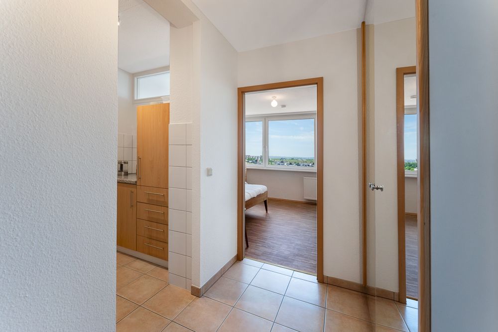 Flur mit Fliesenboden, Blick in Küche und helles Zimmer mit großen Fenstern.