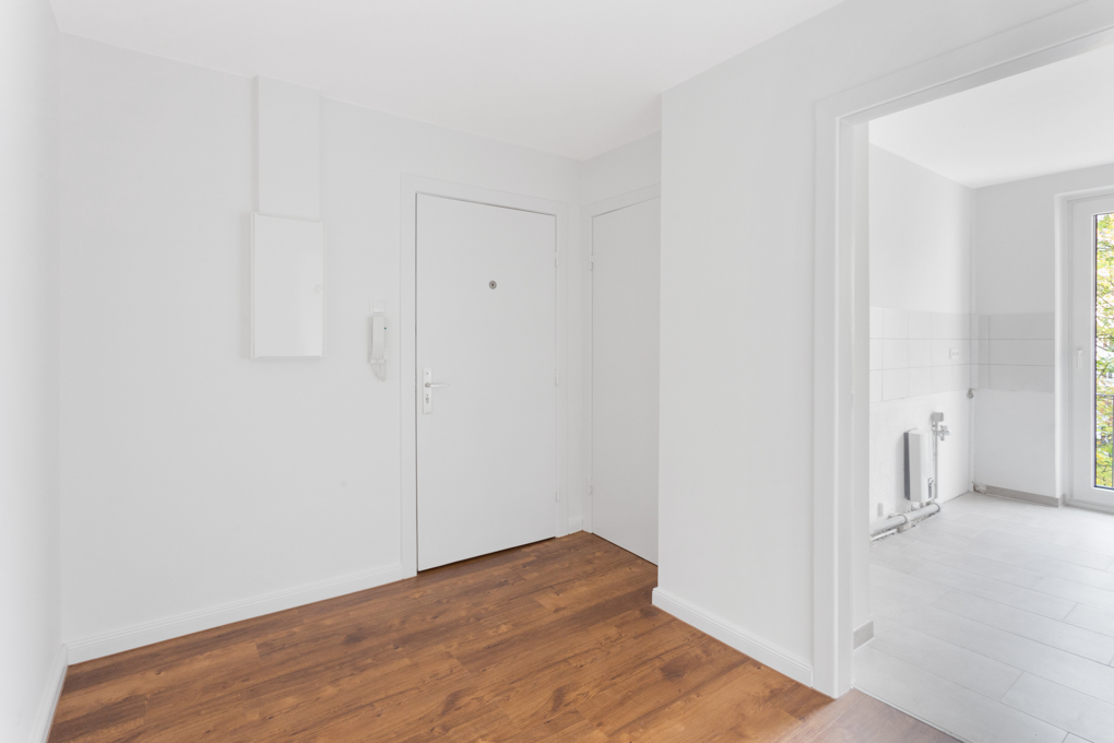 Heller Flur mit Holzboden, weißer Tür und Blick in ein weiteres Zimmer mit Fliesenboden.