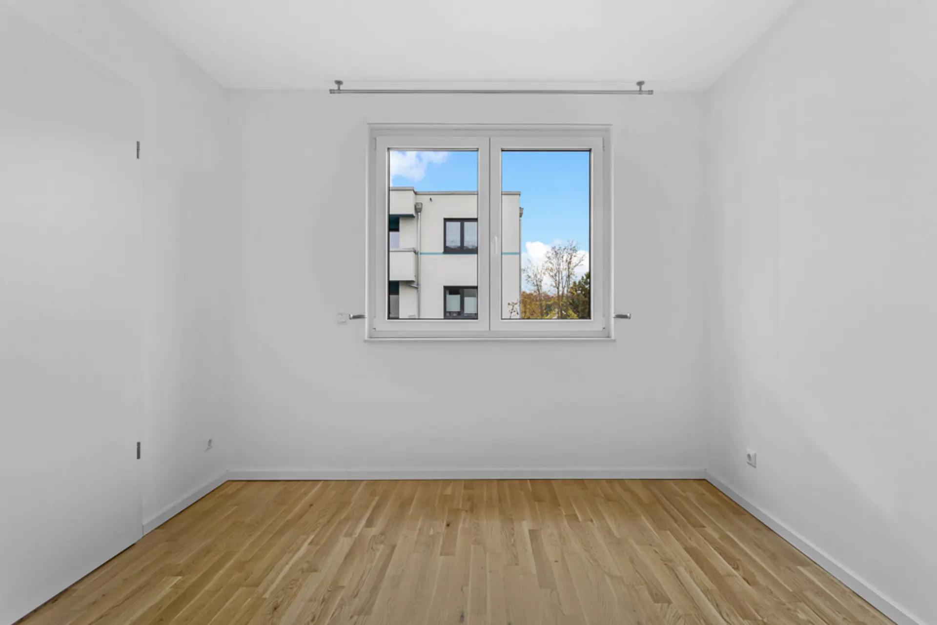Leerer Raum mit Holzboden und Fenster, durch das man ein Nachbargebäude sieht.
