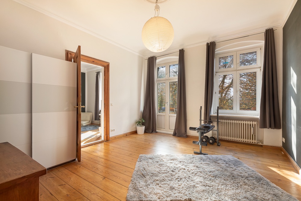 Helles Zimmer mit Holzboden, Teppich, großen Fenstern, Vorhängen und einer Stehlampe.