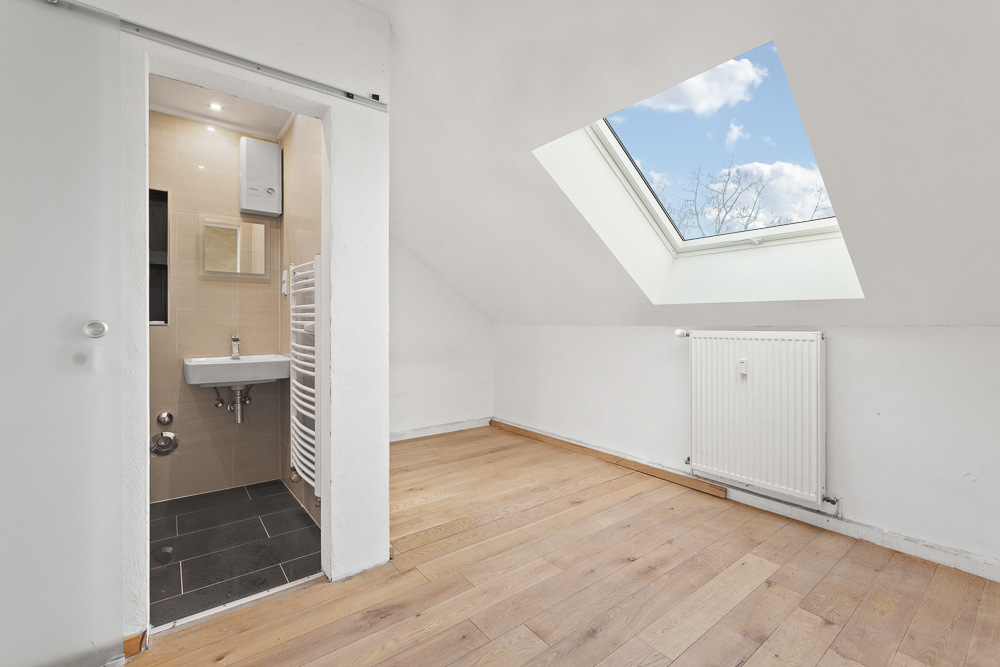 Helles Zimmer mit Dachfenster, Holzfußboden und kleinem Badezimmer mit Waschbecken und Heizkörper.
