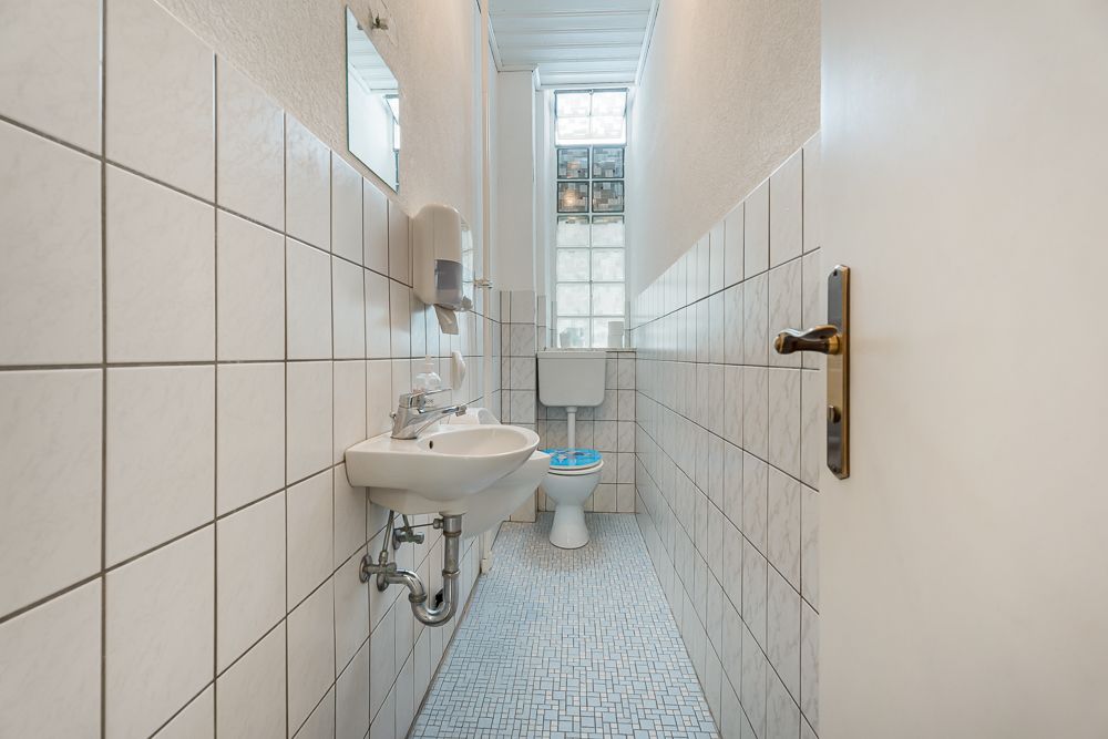 Schmales Badezimmer mit WC, Waschbecken, Spiegel und Fenster mit Glasbausteinen.
