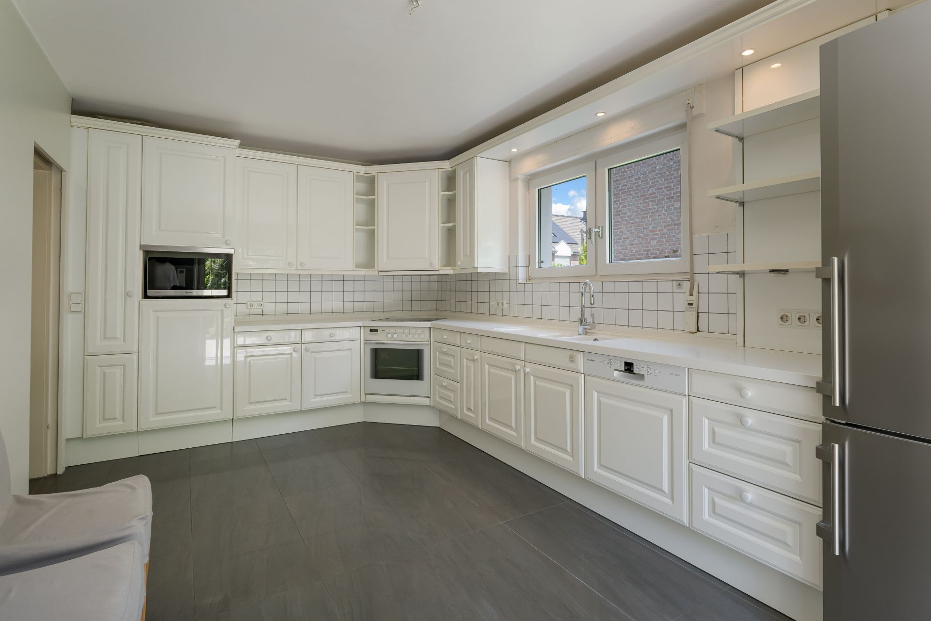 Moderne weiße Einbauküche mit Küchengeräten, Fenstern und grauem Boden.