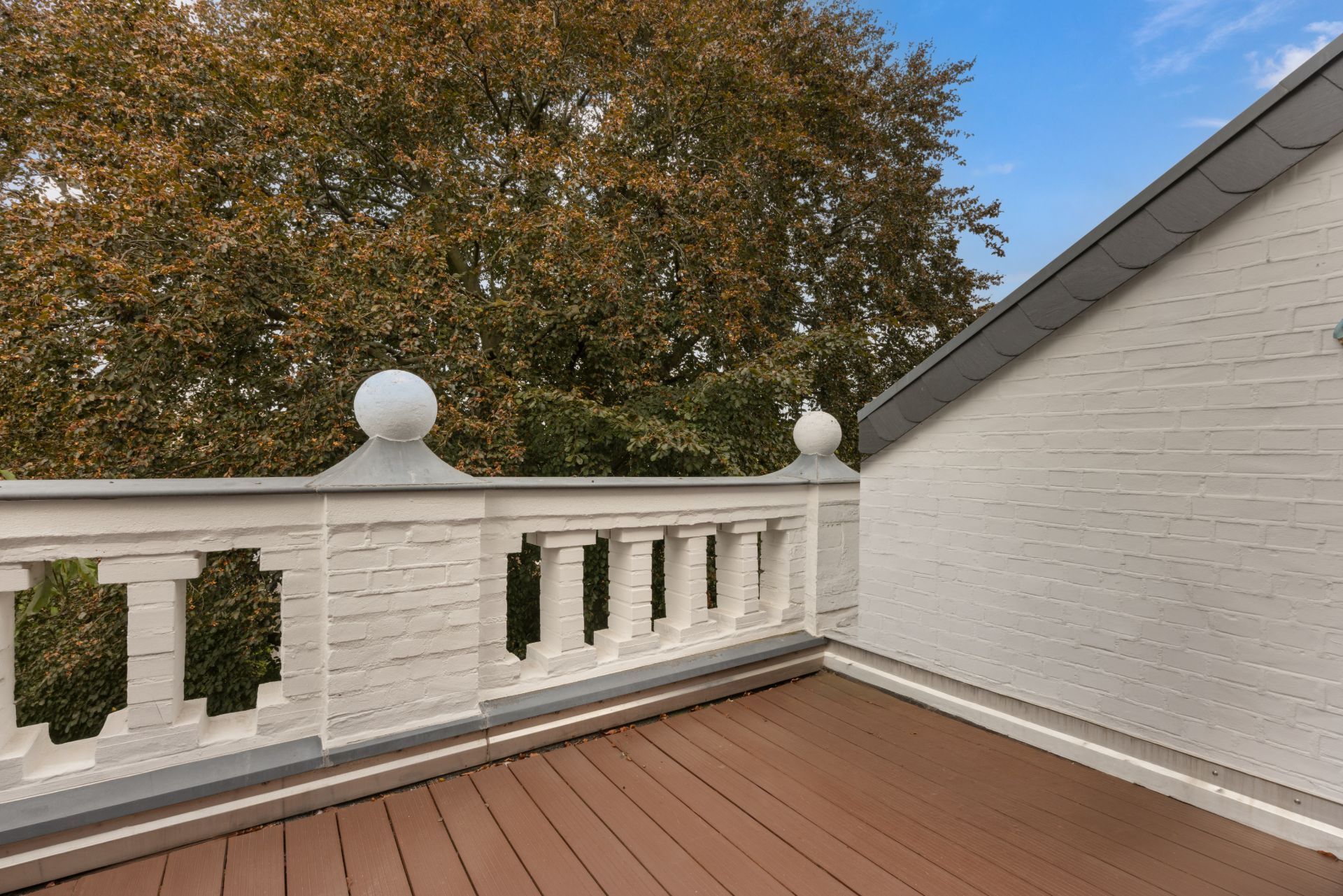 Balkon mit weißem Geländer und braunem Boden, daneben Hauswand, im Hintergrund Baum.