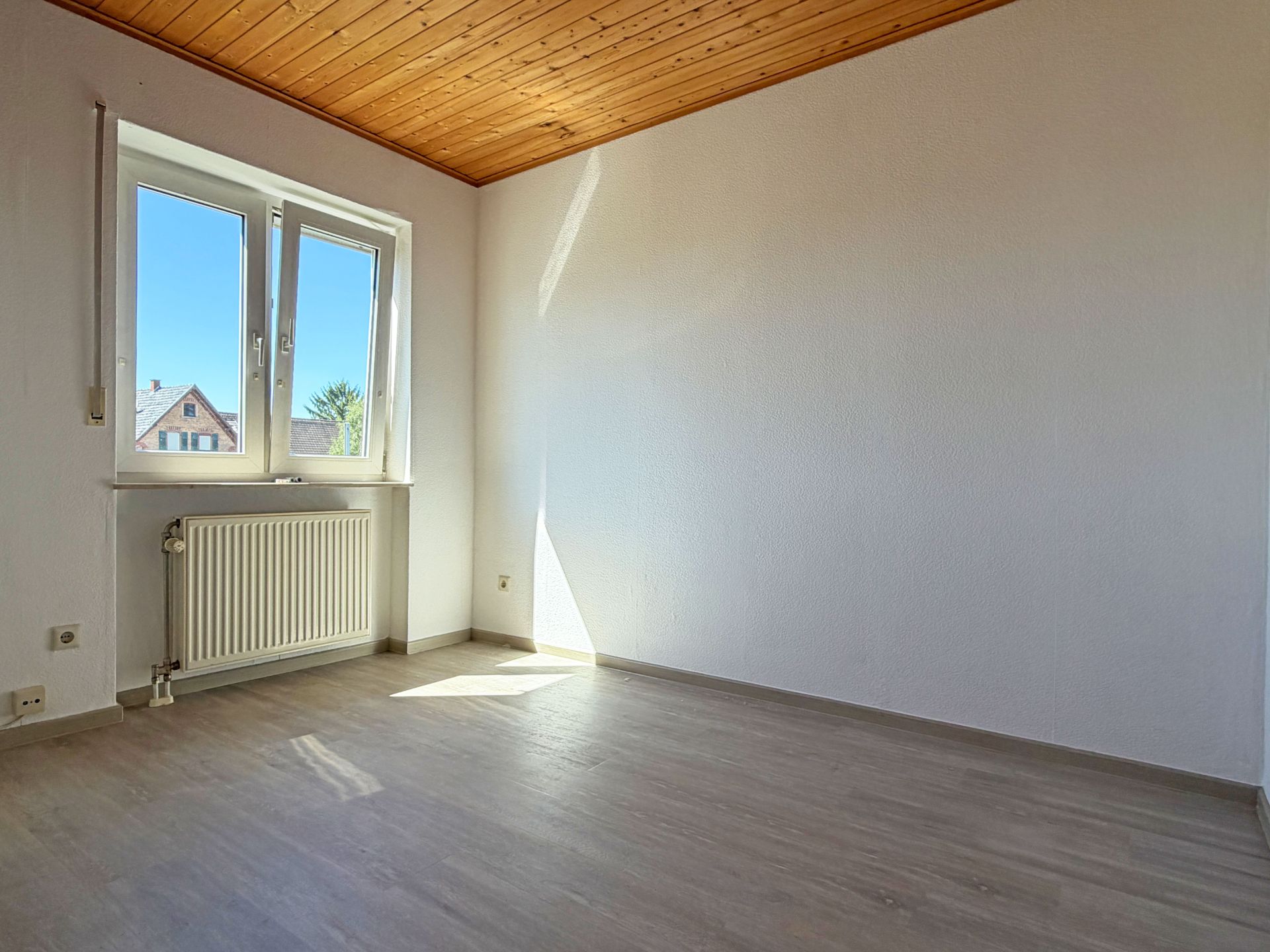 Leerer, heller Raum mit Holzdecke, großem Fenster und Heizkörper unter dem Fenster.