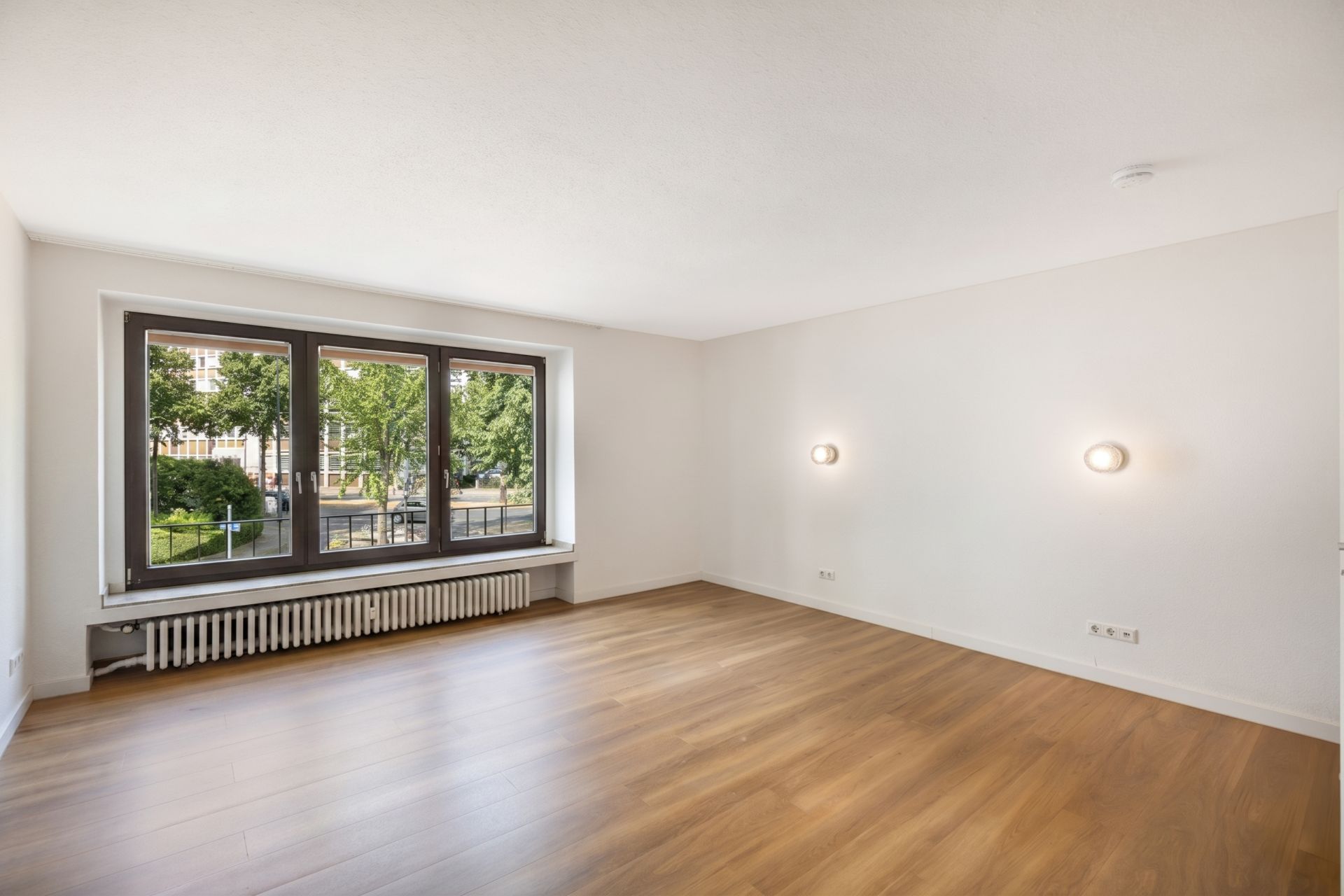 Helles, leeres Zimmer mit großem Fenster, Holzfußboden und zwei Wandleuchten.