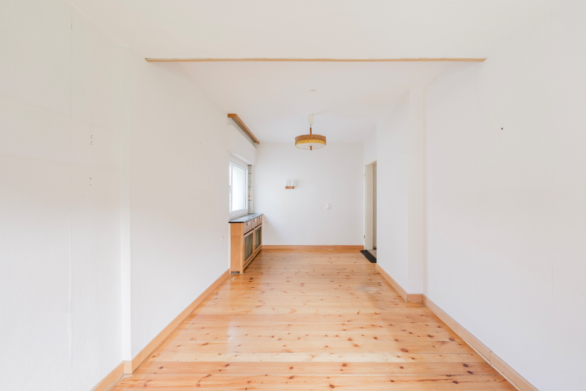 Helles, leeres Zimmer mit Holzboden, weiß gestrichenen Wänden und einem Fenster auf der linken Seite.