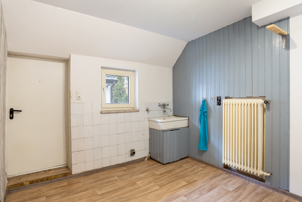 Raum mit kleinem Fenster, Waschbecken, Heizkörper und hellgrauen Wandpaneelen.
