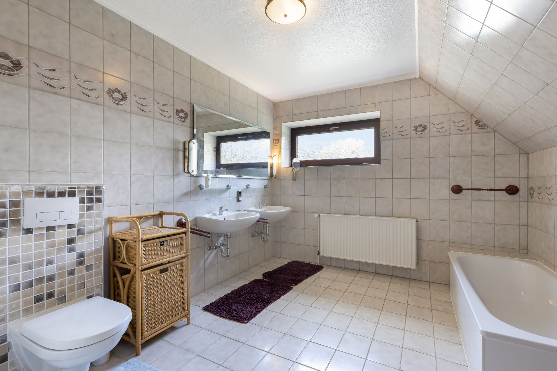 Helles Badezimmer mit Badewanne, zwei Waschbecken, WC, Spiegel und Fenster unter einer Dachschräge.