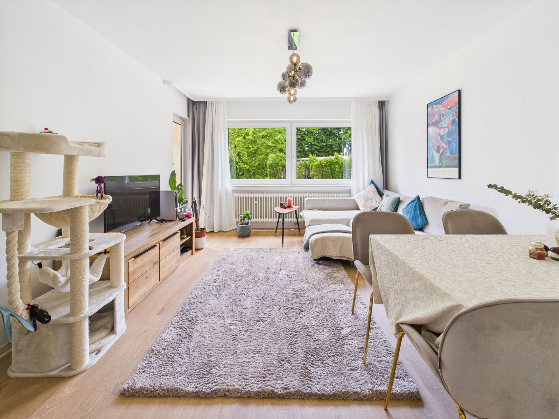 Helles Wohnzimmer mit Sofa, Teppich, Esstisch, Fernseher, Kratzbaum und großen Fenstern.