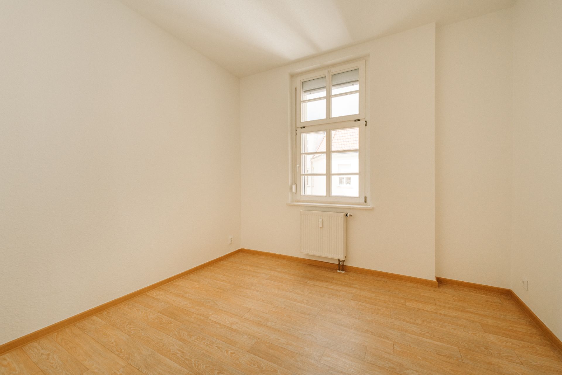 Leerer Raum mit hellem Holzboden und einem Fenster mit Heizkörper darunter.