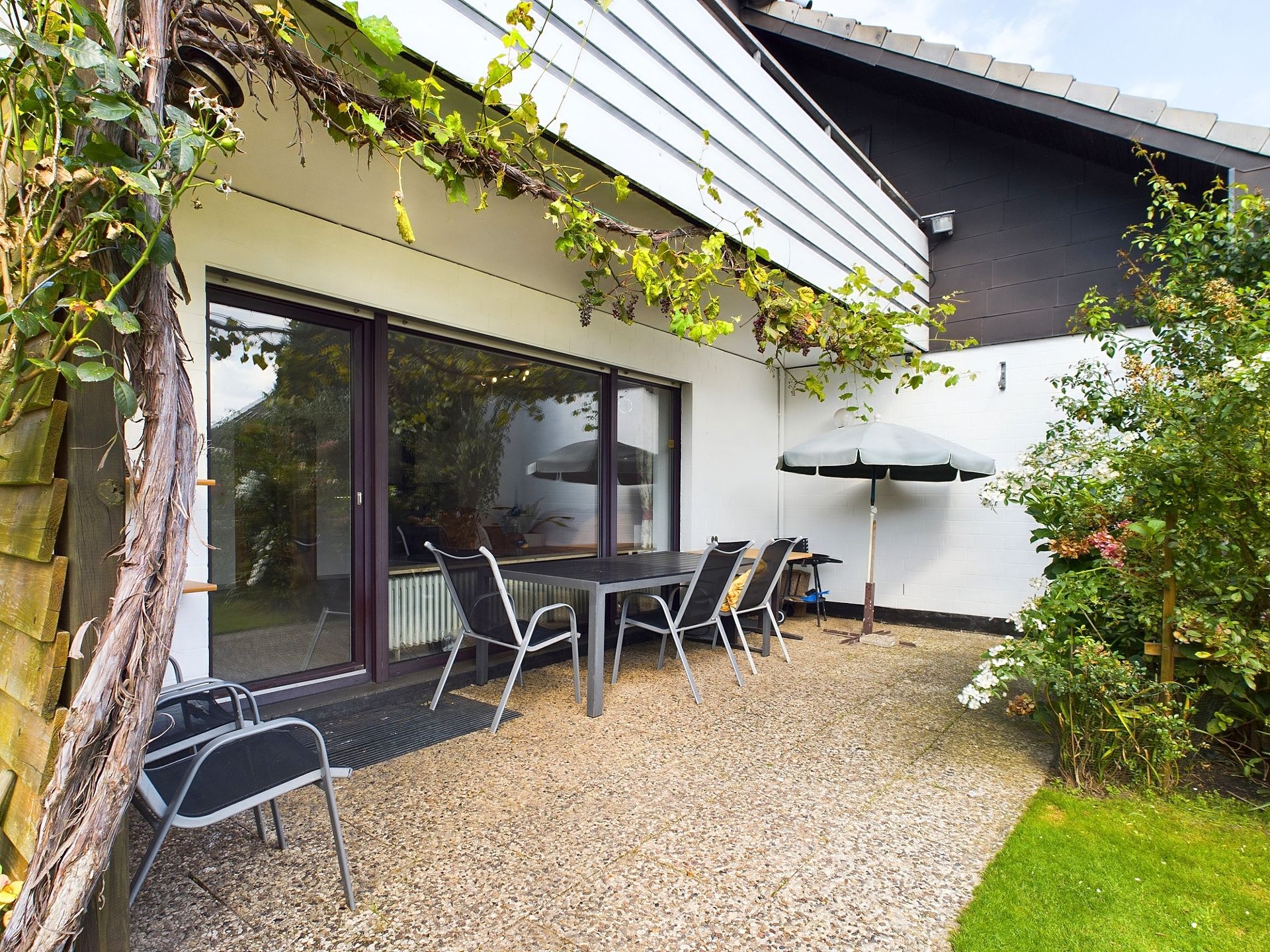 Terrasse mit Tisch, Stühlen, Sonnenschirm und umgeben von Pflanzen neben einem Wohnhaus.