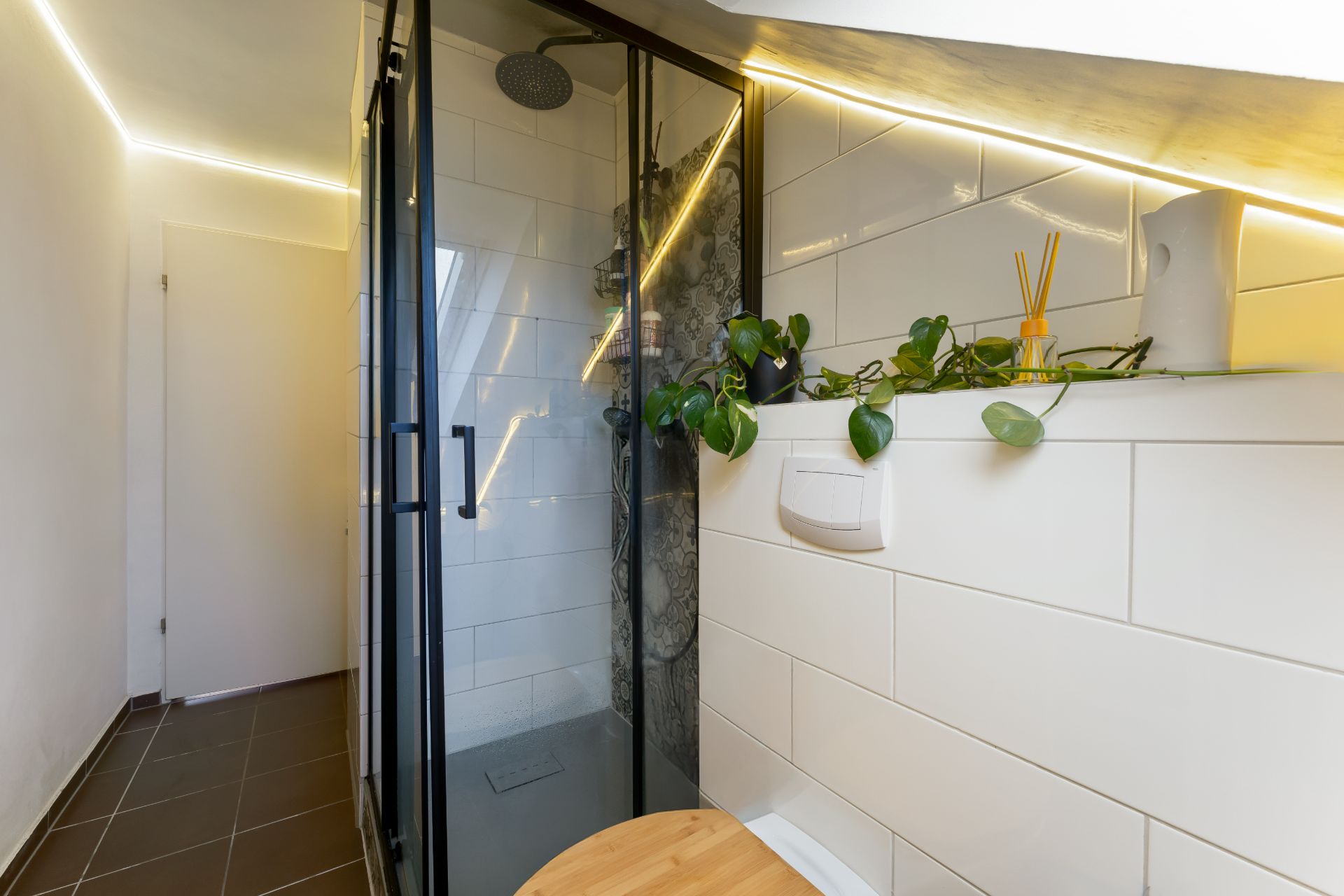 Modernes Badezimmer mit Dusche, weißen Fliesen und einer Pflanze auf einem Regal.
