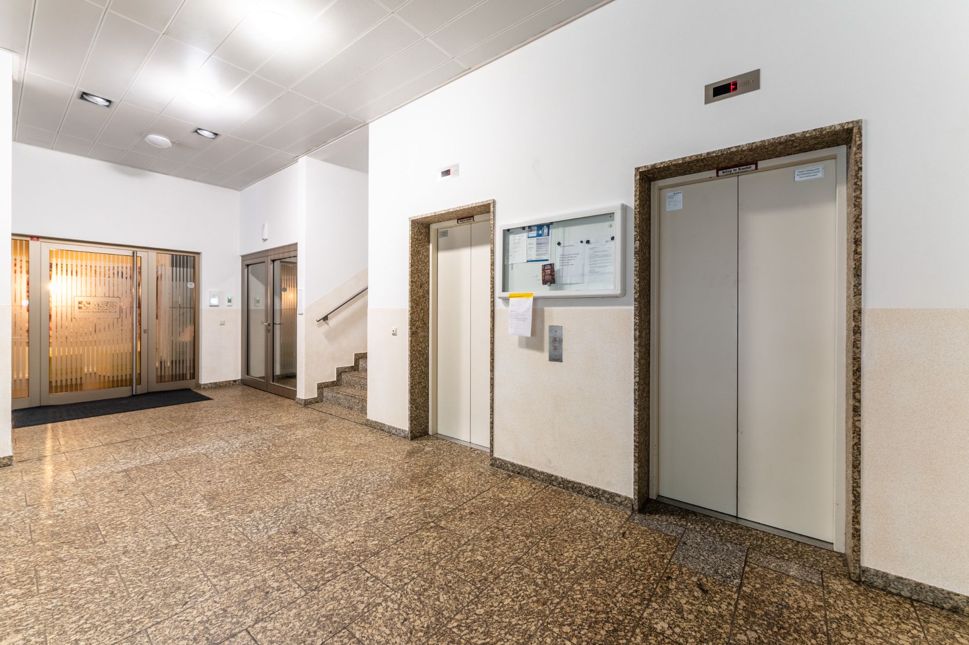 Heller Hausflur mit zwei Aufzügen, Treppe und Glastüren im Hintergrund.