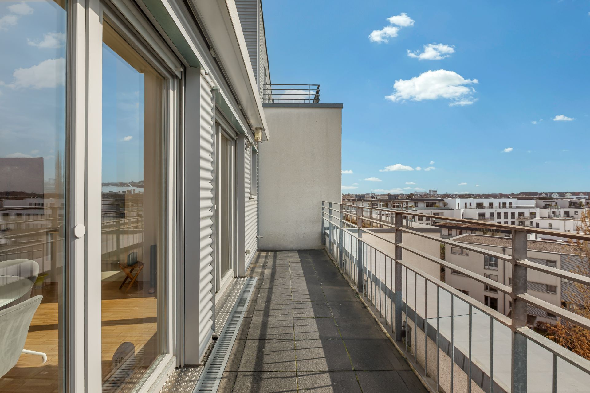 Langer Balkon mit Metallgeländer und Blick auf benachbarte Wohngebäude bei blauem Himmel.