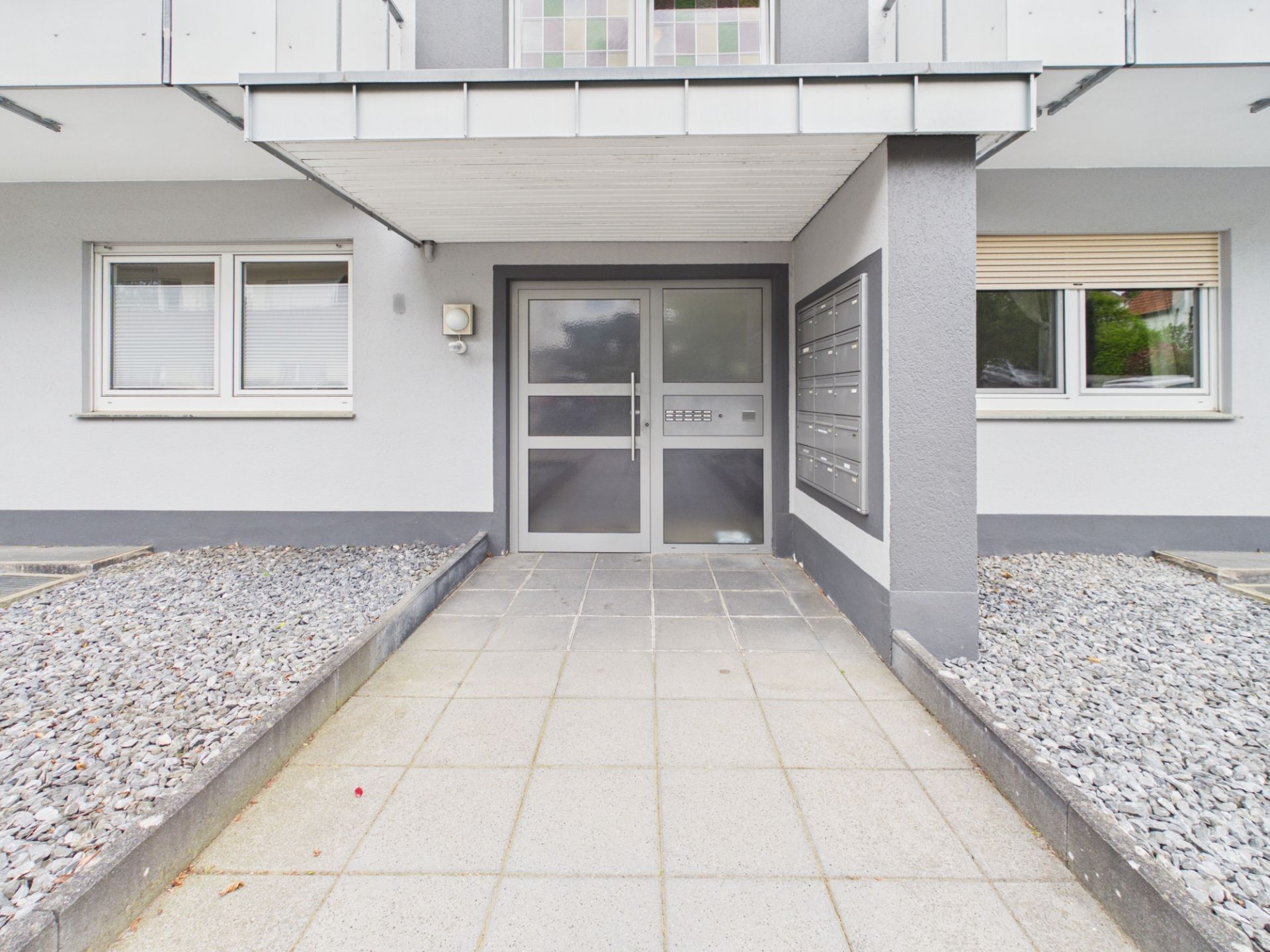 Eingangsbereich eines Wohnhauses mit Glastür, Briefkästen und zwei Fenstern.